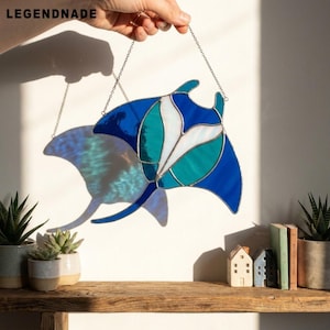 Puede incluir: Un atrapasoles de vidrieras con forma de mantarraya, que presenta segmentos de vidrio azul profundo, verde azulado y blanco unidos por líneas de soldadura de metal oscuro. La pieza está suspendida por una fina cadena de metal.