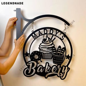 May include: Black metal bakery sign with the text "Maddy's Bakery" and images of a cupcake, donut, and bread. The sign is hanging from a curved metal bracket.