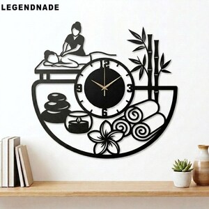 Op de afbeelding: Zwarte wandklok met een spa-thema. De klok heeft een silhouet van een persoon die een massage krijgt, een wijzerplaat, bamboe, handdoeken, stenen, een kaars en een bloem. De wijzers van de klok zijn goudkleurig.
