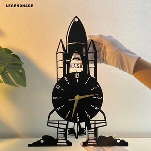 Op de afbeelding: Zwarte wandklok in de vorm van een spaceshuttle, met het NASA-logo en een wijzerplaat. De klok heeft goudkleurige wijzers en een zwart frame. De klok wordt vastgehouden door een persoon met witte handschoenen. Afmetingen in centimeters.