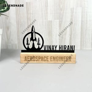 May include: A black desk nameplate with a rocket ship design and the name "VINAY HIRANI". The nameplate sits on a wooden base with the words "AEROSPACE ENGINEER". The design includes three rockets launching.