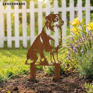 Peut inclure: Une décoration de jardin en métal couleur rouille représentant la silhouette découpée d'un chien assis. Le design inclut le texte LEGENDNADE en haut à gauche. La figurine en métal est plantée dans le sol avec une finition texturée.