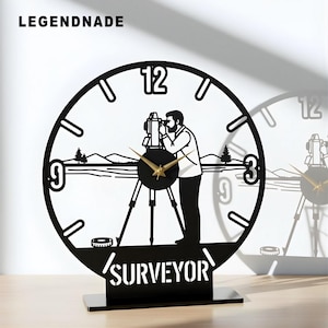 May include: Black metal surveyor clock with a circular design. The clock features a silhouette of a surveyor using a theodolite, with the word "SURVEYOR" at the base. The clock hands are gold-colored, and the brand name "LEGENDNADE" is at the top.