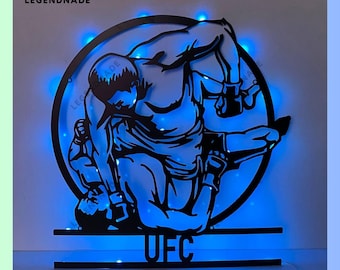 Może przedstawiać: Czarna metalowa ozdoba ścienna przedstawiająca dwie postacie w walce wrestlingowej, z napisem "UFC" poniżej. Ozdoba jest podświetlona niebieskimi diodami LED.