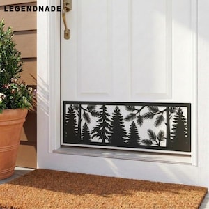 May include: A rectangular black metal kick plate featuring a laser-cut silhouette of a forest with pine trees and branches. The decorative panel is mounted horizontally on the lower section of a white door with a gold handle and text.