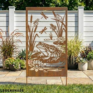 Op de afbeelding: Een roestbruin metalen tuinscherm met een vijvertafereel met een kikker, libellen, waterlelies en riet. Het scherm is rechthoekig met een decoratief frame en twee poten voor plaatsing op de grond. Het woord "LEGENDNADE" staat onderaan.