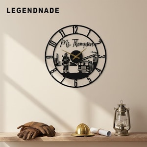 Puede incluir: Reloj de pared de metal negro con temática de bombero. El reloj presenta una silueta de bombero, un camión de bomberos y el texto "Ms. Thompson". Las manecillas del reloj son doradas. La marca "LEGENDNADE" está en la parte superior izquierda.