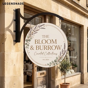 Puede incluir: Un letrero colgante redondo de color crema con un soporte de metal negro. El letrero presenta el texto THE BLOOM & BURROW, Curated Collections y est. 2024, decorado con ilustraciones de ramas de lavanda, hojas de eucalipto y perlas.