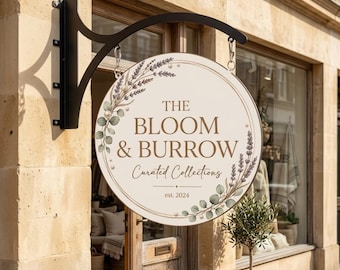 May include: A round, cream-colored hanging sign with a black metal bracket. The sign features the text THE BLOOM & BURROW Curated Collections est. 2024, decorated with illustrations of lavender sprigs, eucalyptus leaves, and small white pearls.