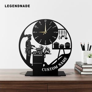 May include: Black metal clock with a chef design. The clock features a chef cooking, a clock face, and the words "CUSTOM NAME". The clock is on a black stand and the brand name "LEGENDNADE" is at the top left.