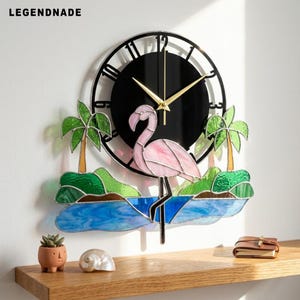 Könnte beinhalten: Eine runde Wanduhr mit schwarzem Zifferblatt, goldenen Zeigern und Ziffern. Unter der Uhr befindet sich ein Buntglas-Motiv, das einen rosa Flamingo im blauen Wasser zwischen zwei grünen Palmen auf kleinen Inseln zeigt. Der Text lautet LEGENDNADE.