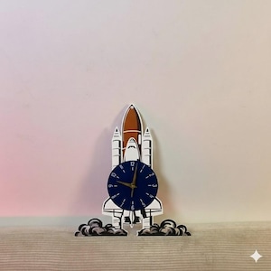 Op de afbeelding: Een decoratieve klok in de vorm van een spaceshuttle, met een blauwe wijzerplaat en gouden wijzers. De shuttle is wit, bruin en zwart, met rook aan de basis. Een uniek, ruimtethema-decoratie-item.