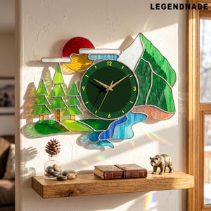 May include: A stained glass wall clock with a nature scene. The clock face is dark green with gold hands and numbers. The scene includes a mountain, trees, a river, and a red sun. The clock is mounted on a white wall above a wooden shelf.