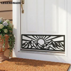 May include: A rectangular black metal door kick plate featuring a laser-cut design of a mountain range beneath a rising sun with radiating beams. The decorative panel is mounted horizontally on the lower section of a white paneled door.