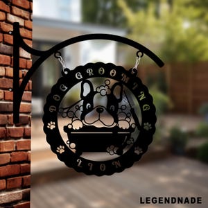 Puede incluir: Letrero de peluquería canina de metal negro con diseño de bulldog francés. El letrero presenta la palabra "GROOMING" en un patrón circular, un perro en una bañera, huellas de patas y un peine. Cuelga de un soporte de metal negro.