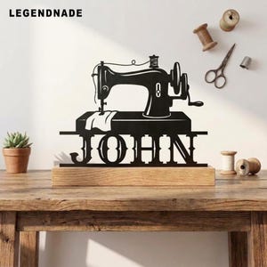 Könnte beinhalten: Eine schwarze Metallsilhouette einer Vintage-Nähmaschine auf einem rechteckigen Holzsockel. Das Design zeigt den Namen JOHN in großen Großbuchstaben, die in die Basis integriert sind. Das Stück wird mit Nähzubehör in der Nähe präsentiert.