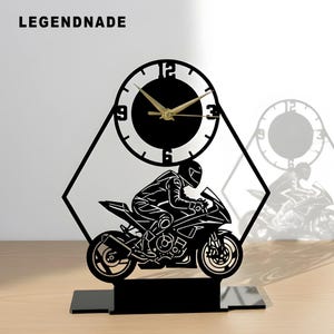 Könnte beinhalten: Schwarze, lasergeschnittene Schreibtischuhr mit Motorradrennfahrer-Design. Die Uhr hat ein rundes Zifferblatt mit goldenen Zeigern und schwarzem Hintergrund. Der Sockel ist rechteckig, und die Uhr ist von einer geometrischen Form umrahmt. Der Text "LEGENDNADE" steht oben.