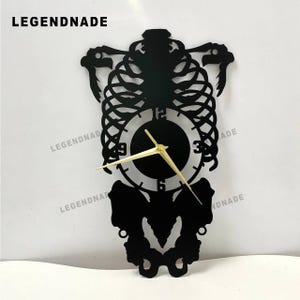 May include: Black metal wall clock shaped like a human rib cage with a circular clock face. The clock hands are gold, and the numbers are visible on the clock face. The brand name "LEGENDNADE" is at the top.
