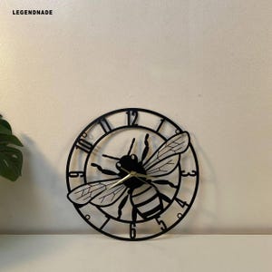 May include: Black metal wall clock featuring a bee design. The clock has a circular frame with Roman numerals and a bee silhouette in the center. The clock hands are gold. The brand name "LEGENDNADE" is in the top left corner.