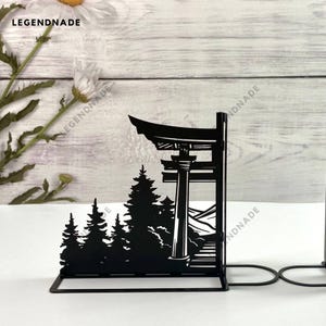 May include: Black metal bookends shaped like a Japanese torii gate with trees and mountains in the background.