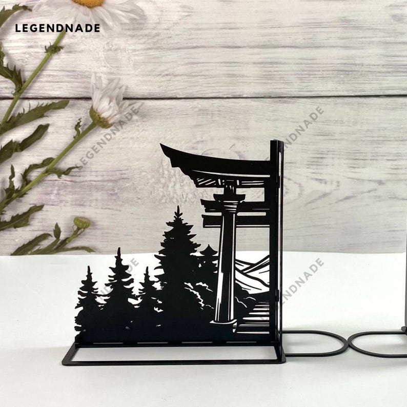 May include: Black metal bookends shaped like a Japanese torii gate with trees and mountains in the background.
