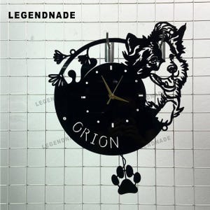 Puede incluir: Reloj de pared negro con diseño de perro, el nombre "ORION" y una huella de pata. El reloj tiene manecillas doradas y la marca "LEGENDNADE". Cuelga de una rejilla blanca. Aproximadamente 25 cm de diámetro.