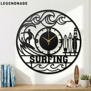 Könnte beinhalten: Schwarze Metall-Wanduhr mit Surf-Motiv. Die Uhr zeigt einen Surfer, Wellen, Surfbretter und einen Leuchtturm. Das Wort "SURFING" ist prominent dargestellt. Die Uhrzeiger sind goldfarben.