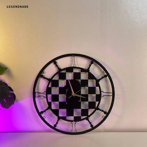 May include: Black and white chess-themed wall clock. The clock face features a checkered pattern, with gold-colored hands. The outer ring has a decorative design. The word "LEGENDNADE" is in the upper left corner.