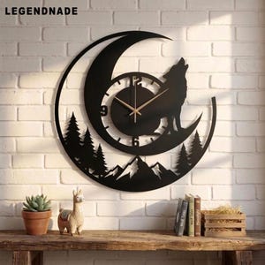 Könnte beinhalten: Eine schwarze Wanduhr in Form einer Mondsichel mit der Silhouette eines heulenden Wolfes, Tannenbäumen und Berggipfeln. Das Zifferblatt hat eine kreisförmige Mitte mit weißen Zahlen und dünnen goldenen Zeigern. Der Text LEGENDNADE ist sichtbar.