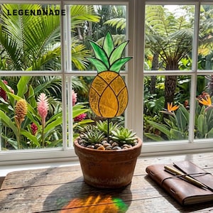 May include: A stained glass pineapple ornament with a yellow body and green leaves, mounted on a thin black metal stake. The ornament is inserted into a terracotta pot filled with small green succulents and pebbles, resting on a wooden surface.