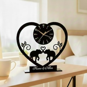 Può includere: Orologio decorativo nero con cornice a forma di cuore. L'orologio ha un quadrante circolare con lancette e numeri. Due sagome di elefanti si fronteggiano sotto l'orologio. La base recita "Hana & Allen".