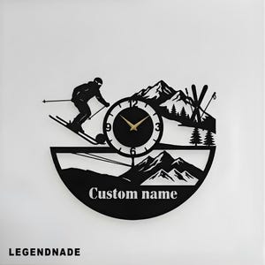May include: Black metal wall clock depicting a skier, mountains, and skis. The clock face has gold hands and a custom name below. The clock is a decorative item with a winter sports theme.