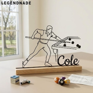 Puede incluir: Una escultura de metal negro de una persona jugando al billar, montada sobre una base de madera. El nombre "Cole" está escrito en cursiva. La escultura está junto a bolas de billar, tiza y un afilador de puntas. La palabra "LEGENDNADE" está en la esquina superior izquierda.