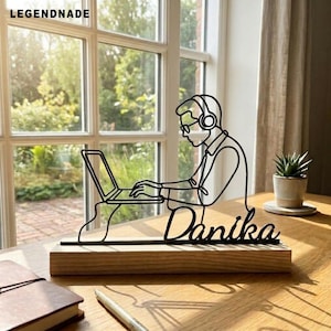 May include: A black wire art sculpture depicting a person wearing headphones and working on a laptop, mounted on a rectangular wooden base. The name Danika is written in cursive script at the bottom of the wire figure, which sits on a desk.
