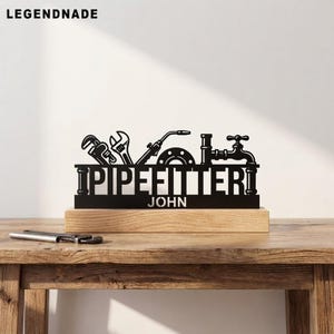 以下が含まれることがあります： PIPEFITTERという文字とJOHNという名前が刻まれた黒い金属製の看板。パイプレンチ、モンキーレンチ、溶接トーチ、蛇口のシルエットが上部にデザインされています。看板は明るい色の長方形の木製ベースに取り付けられています。
