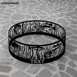 May include: Black metal fire pit ring with a cut-out design of fish and aquatic plants. The word "TEXT" is displayed at the bottom. The fire pit ring is circular and sits on a stone surface. The brand name "LEGENDNADE" is at the top.