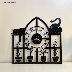 May include: Black metal wall clock featuring a cat design. The clock has Roman numerals and a black face with gold hands. The clock also includes a window design with potted plants and two cat silhouettes. The brand name "LEGENDNADE" is in the top left corner.