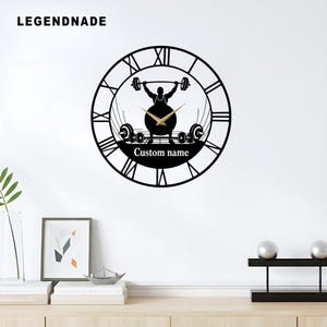May include: Black wall clock with a weightlifter silhouette lifting a barbell. The clock features Roman numerals and gold-colored clock hands. The text "Custom name" is displayed below the weightlifter.
