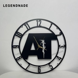 May include: Black metal wall clock with a circular design. The clock features a cutout design with the letters "AT" in a rectangular shape at the centre. Gold-coloured clock hands and the numbers 1 through 12 are visible. The brand name "LEGENDNADE" is at the top.