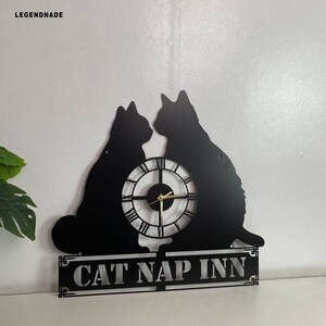 Puede incluir: Reloj de pared de metal negro con dos siluetas de gatos, una esfera con n&uacute;meros romanos y el texto "CAT NAP INN". El reloj tiene manecillas doradas y est&aacute; dise&ntilde;ado para montaje en pared.