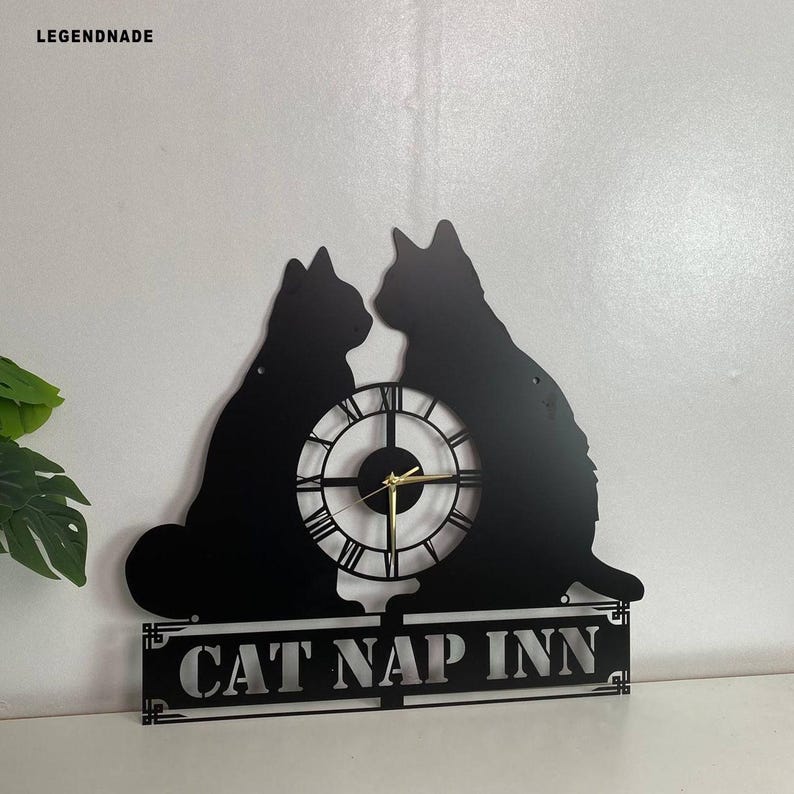 Puede incluir: Reloj de pared de metal negro con dos siluetas de gatos, una esfera con n&uacute;meros romanos y el texto "CAT NAP INN". El reloj tiene manecillas doradas y est&aacute; dise&ntilde;ado para montaje en pared.