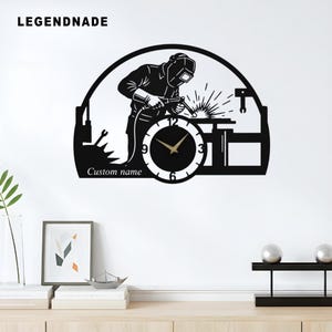 Peut inclure: Horloge murale en métal noir représentant un soudeur en silhouette, avec des étincelles. Le cadran de l'horloge est blanc avec des chiffres noirs et des aiguilles dorées. Le texte "Custom name" est en dessous. Le mot "LEGENDNADE" est en haut à gauche.