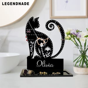 Peut inclure: Organisateur de bijoux noir en forme de chat avec une base et une plaque signalétique avec le nom "Olivia". L'organisateur comporte des trous pour les boucles d'oreilles et une queue incurvée pour les colliers. La base contient des bagues et une montre.