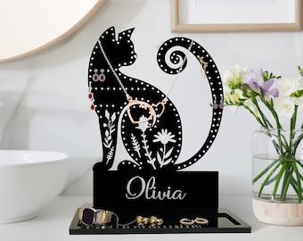 Pode incluir: Organizador de joias preto em forma de gato com base e placa de identificação com o nome "Olivia". O organizador tem orifícios para brincos e uma cauda curva para colares. A base contém anéis e um relógio. O logotipo Legendnade está no canto superior esquerdo.