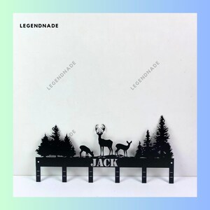 May include: Black metal coat rack with a silhouette of a forest scene, including deer and trees. The rack has four hooks and the name "JACK" is written below the scene.