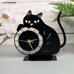 May include: Black cat-shaped clock with gold hands and a round clock face. The cat is posed as if it is holding the clock. The clock is on a black base. The word "LEGENDNADE" is in the top left corner.