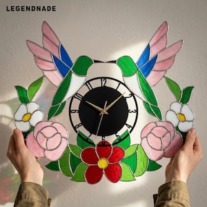 Könnte beinhalten: Eine Wanduhr aus Buntglas mit zwei Kolibris mit grünen, blauen und rosa Flügeln, umgeben von rosa und weißen Blumen mit gelben Mitten und grünen Blättern. Die Uhr hat ein schwarzes Zifferblatt mit goldenen Zeigern und dem Text LEGENDNADE.