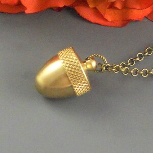 May include: A gold-colored acorn pendant necklace. The acorn pendant has a textured cap and a smooth, rounded body. The chain is a delicate, antique gold color. The background includes a vibrant orange flower.