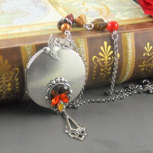 May include: A silver locket necklace with a bird charm and a red, orange, and yellow crystal embellishment. The necklace is on a brown book with gold accents.