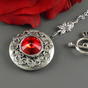 May include: A silver locket necklace featuring a red crystal at its centre. The locket is adorned with a delicate floral design. The necklace has a toggle clasp with a crown detail.